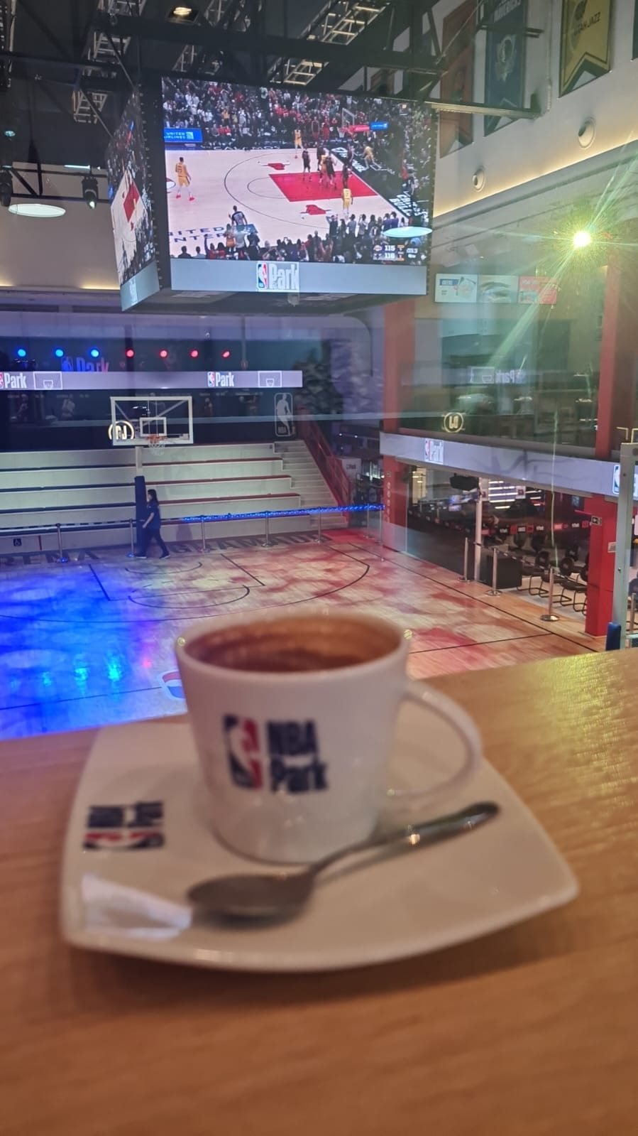 Xícara de café com o logo do NBA Park sobre o balcão, com a quadra de basquete e o telão ao fundo