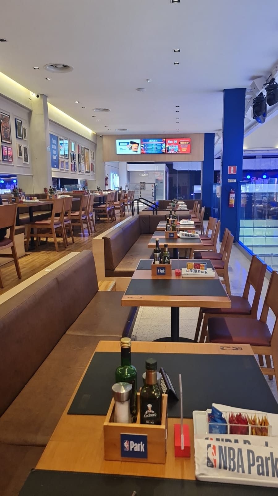 Mesas de madeira e bancos estofados do restaurante do NBA Park, com pilares azuis e telões ao fundo