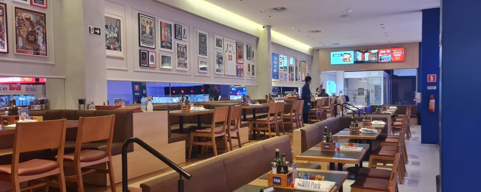 Salão do restaurante do NBA Park, com quadros de basquete nas paredes, mesas de madeira e telões com o cardápio